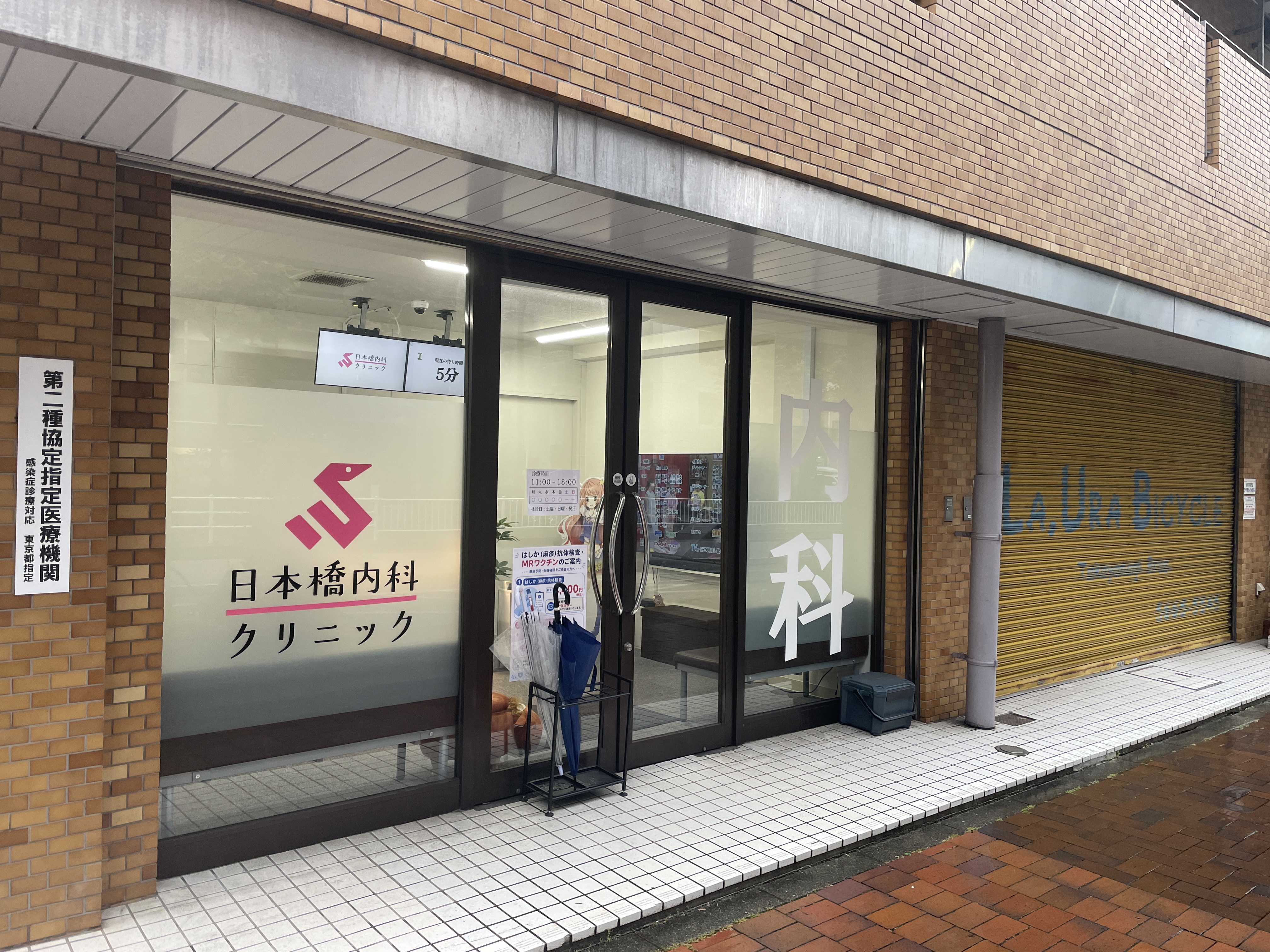日本橋内科クリニック 외관 — 비오는 날의 동네 내과 클리닉 입구. 내과라는 큰 글씨가 유리창에 새겨져 있다